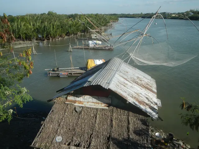The image shows a hut on the shore of a body of water with a fishing net in the foreground. In the...