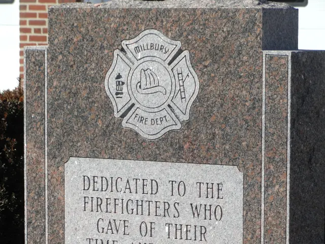 The image shows a memorial stone with a plaque on it in front of a brick building, surrounded by...