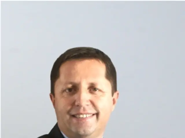 The image shows a man in a suit and tie with his arms crossed, smiling against a white background....
