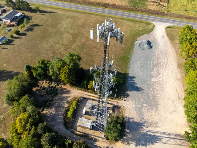 The image shows an aerial view of a cell phone tower in the middle of a field, surrounded by trees,...