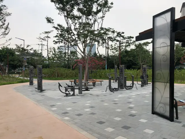The image shows a park with a variety of outdoor fitness equipment, including benches, trees,...