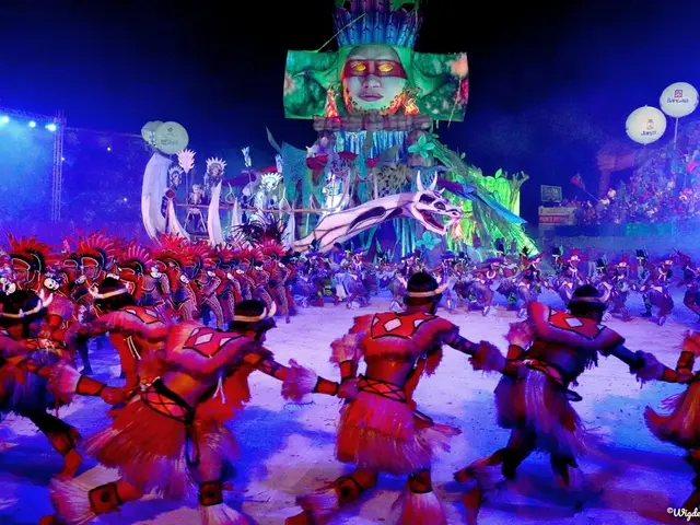 The image shows a group of people in colorful costumes dancing on a stage in front of a crowd of...