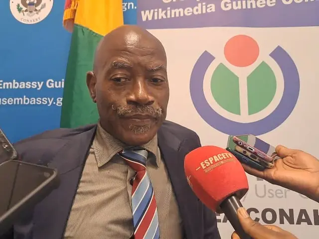 The image shows a man in a suit and tie talking to reporters, with a person's hand holding a...