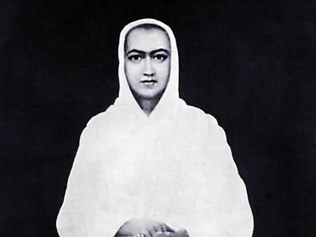 The image shows a black and white photo of a woman sitting in a lotus position on a platform, with...