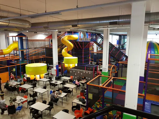 The image shows a large indoor play area with tables and chairs, people sitting on the chairs and...
