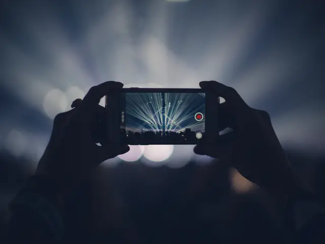 The image shows a person taking a photo of a concert with their cell phone. The person's hands are...