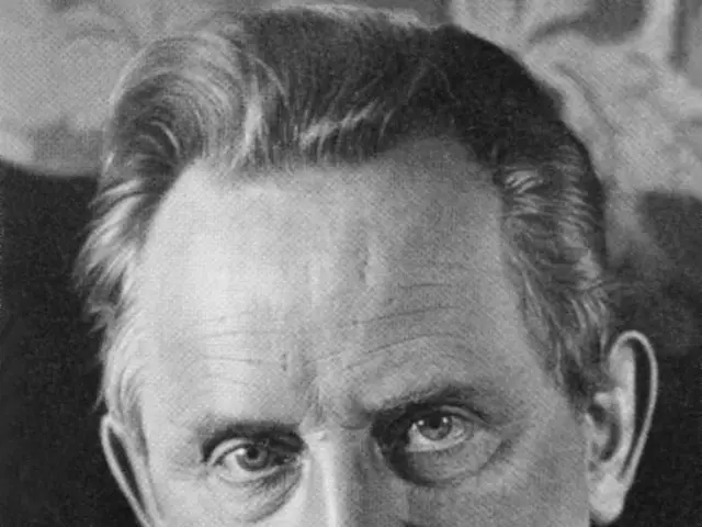 The image shows a black and white photo of a man wearing a blazer and bow tie, with a blurred...