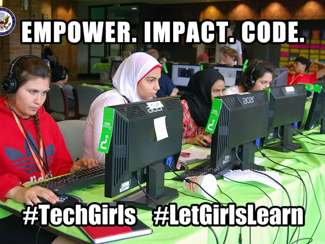 The image shows a group of young women sitting at a table with computers, keyboards, mouses, books,...