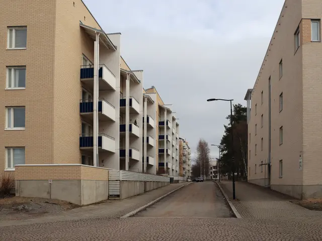The image shows an empty street with tall apartment buildings on either side. The buildings have...