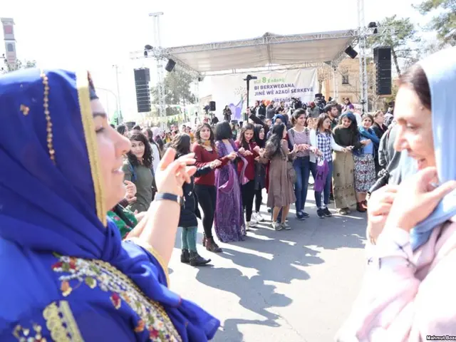 The image shows a group of women standing in front of a crowd of people on a road surrounded by...