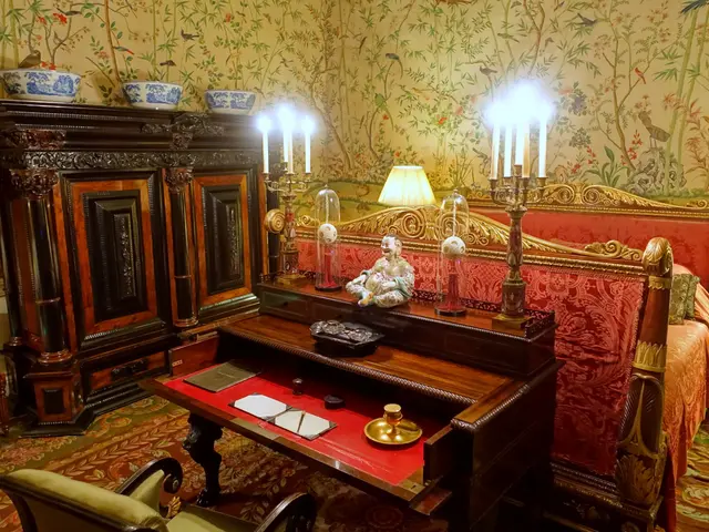 The image shows a bedroom with a bed, a desk, a chair, a table with books, papers, and other...