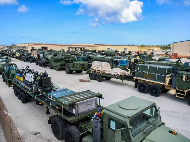 The image shows a long line of military trucks parked in a parking lot, with a mesh fencing on the...