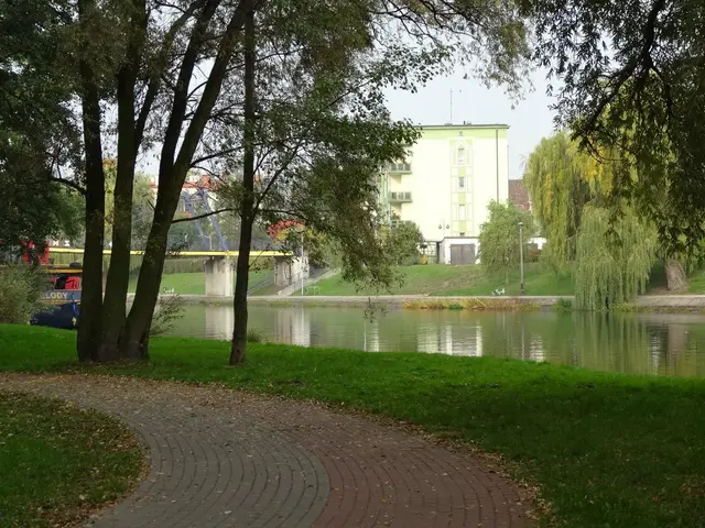 The image shows a path winding through a park next to a body of water, with lush green grass on...