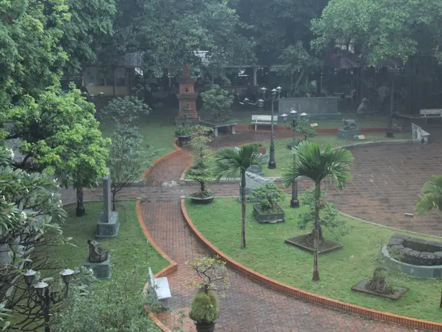 The image shows a lush green park with a variety of trees, plants, grass, light poles, benches, and...