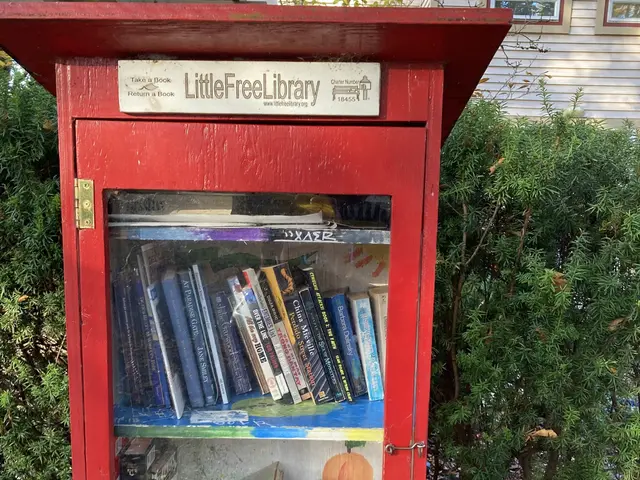 The image shows a red little free library in front of a house, surrounded by plants and trees....