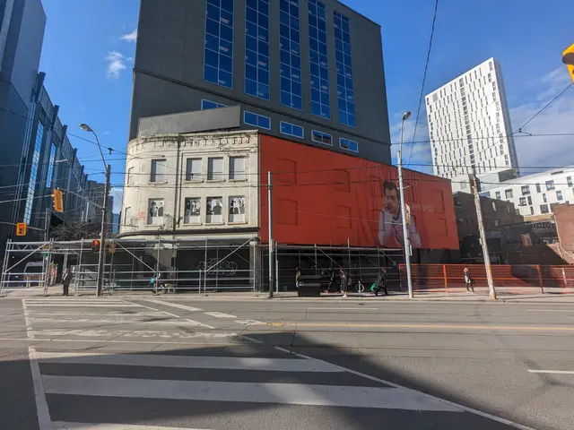The image shows a large building with a large mural on the side of it, surrounded by street poles,...