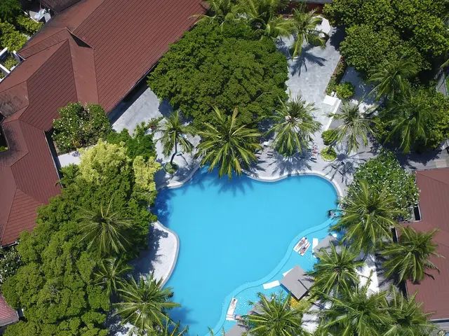 The image shows an aerial view of a resort with a large swimming pool surrounded by lush green...