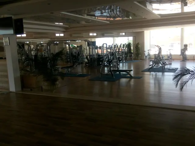 The image shows a large gym with a variety of exercise equipment, houseplants, and a television...