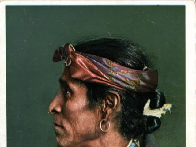 The image shows an old photo of a Native American man wearing a traditional dress and ornaments. At...