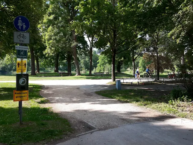 The image shows a park with a paved path in the middle of it, surrounded by trees and grass. On the...