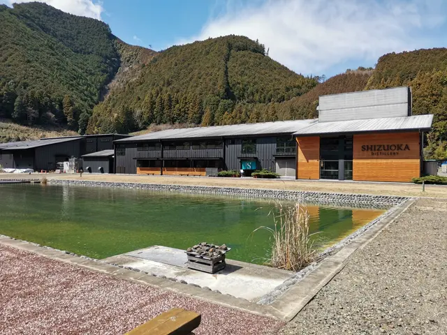 The image shows a large building with a pond in front of it, surrounded by lush green plants and...