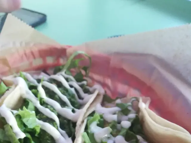 The image shows two fish tacos in a basket on a table, with a person's hand holding a mobile phone...