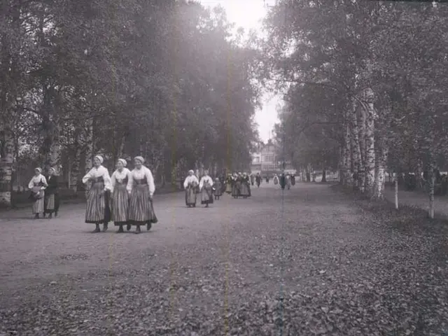 The image shows a group of women walking down a path in a park, surrounded by trees and a building...