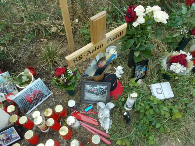 The image shows a memorial in the grass with a cross, candles, flowers, photo frames, and other...