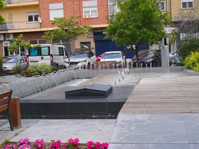 The image shows a fountain in the middle of a city square surrounded by benches, plants, flowers,...