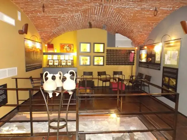 The image shows a room filled with lots of tables and chairs, a railing, vases, boards with text...