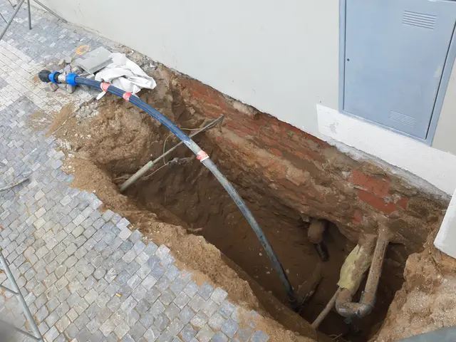 The image shows a large hole in the ground next to a building, with pipes and other objects...