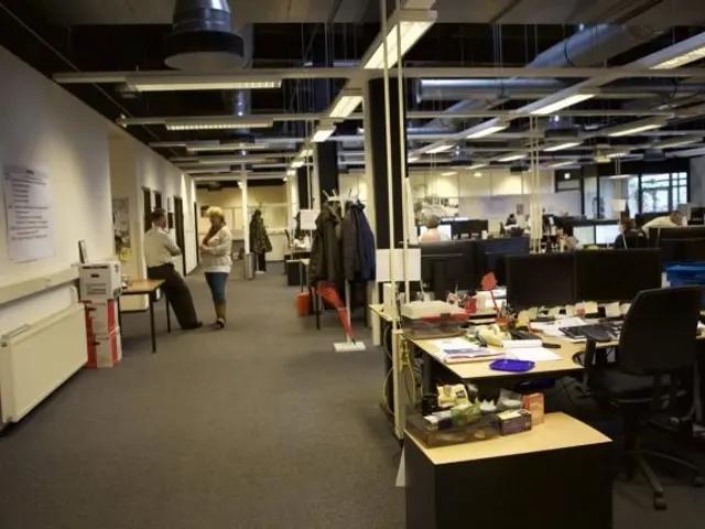 The image shows a large open plan office with people working on computers, desks, chairs, monitors,...