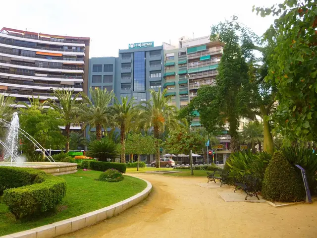 The image shows a park with a fountain in the middle of it surrounded by lush green grass, plants,...