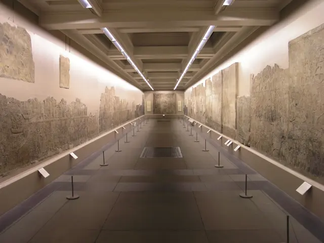 The image shows a long hallway in a museum with walls adorned with carvings, poles with ropes, and...