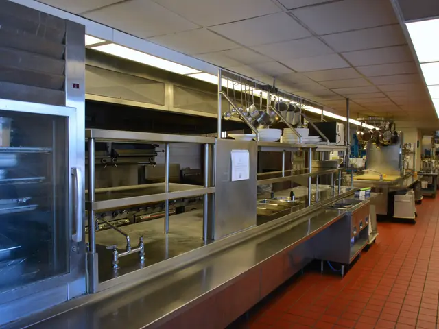The image shows a commercial kitchen with stainless steel counter tops, electric lights to the...
