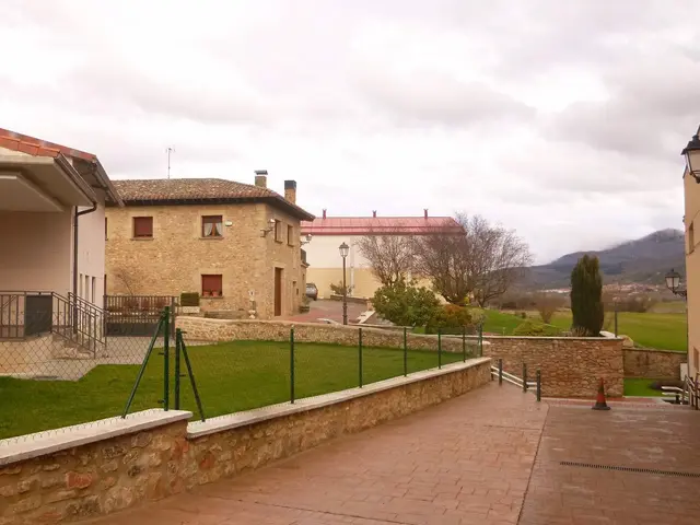 The image shows a building with windows, railings, steps and a roof, surrounded by a metal fence,...