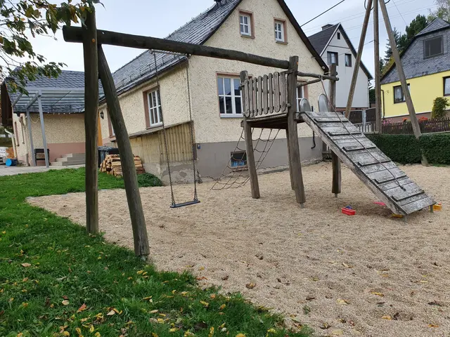 The image shows a playground with a wooden swing set and a slide in the middle of it, surrounded by...