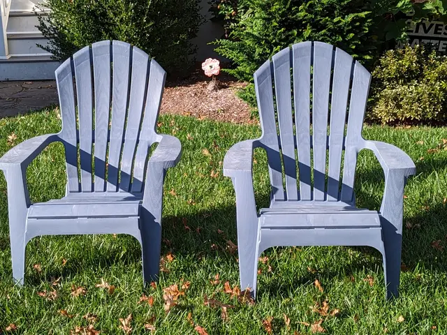 The image shows two blue Adirondack chairs sitting in the grass in front of a house. The chairs are...