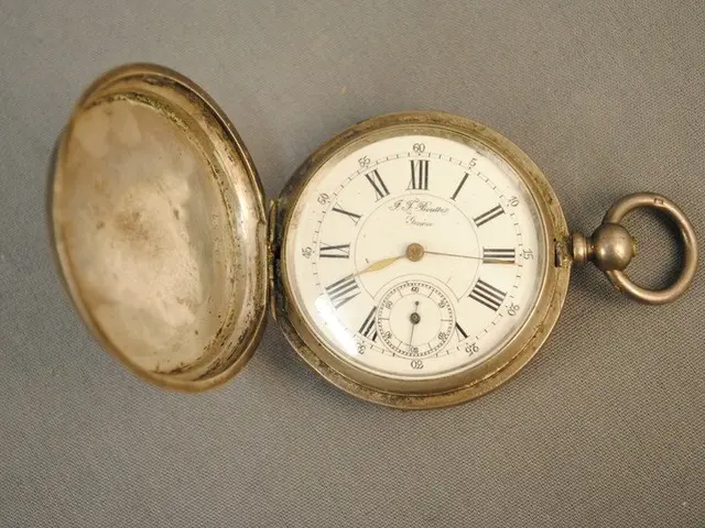 The image shows a silver pocket watch with a white dial featuring Roman numerals on a grey surface....
