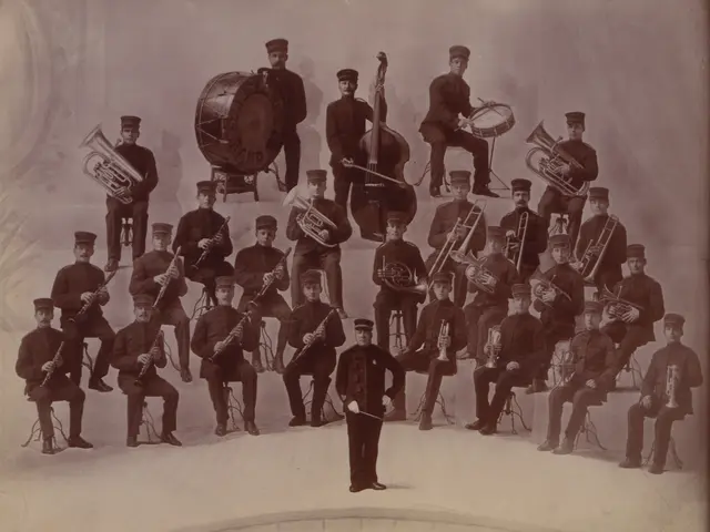 The image shows an old photo of a band posing for a picture. There are a few people sitting and a...