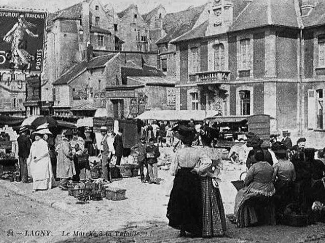 The image shows an old black and white photo of a bustling market in the old town of Ligny, France....