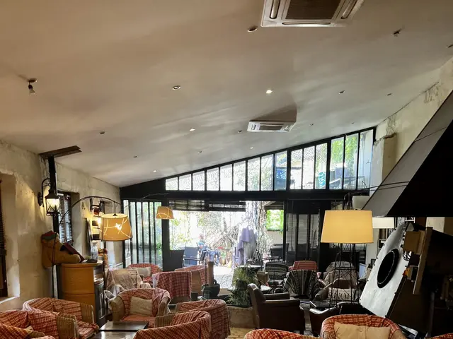 The image shows a large room filled with lots of chairs and tables, cushions on the sofas, lamps on...