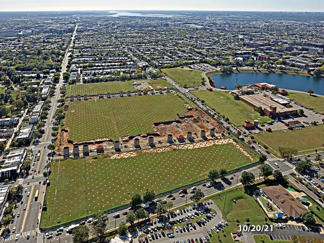 The image shows an aerial view of a city with lots of buildings, trees, grass, vehicles on the...