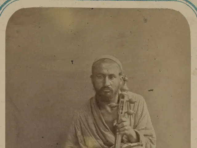 The image shows an old photo of a man sitting on the floor, holding a musical instrument in his...