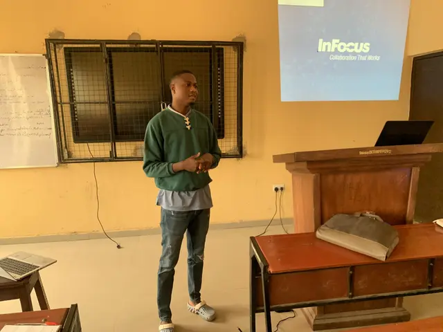 The image shows a man standing in front of a projector screen in a classroom. He is wearing a green...