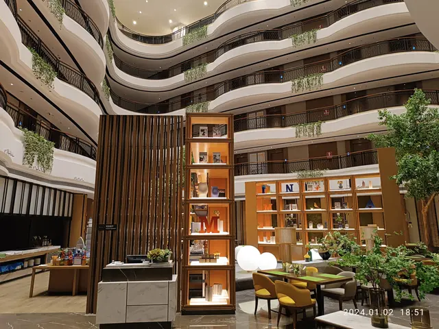 The image shows a large atrium filled with lots of tables and chairs, houseplants, railings,...