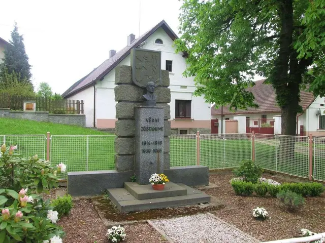 The image shows a monument in the middle of a garden surrounded by plants with flowers, a fence,...