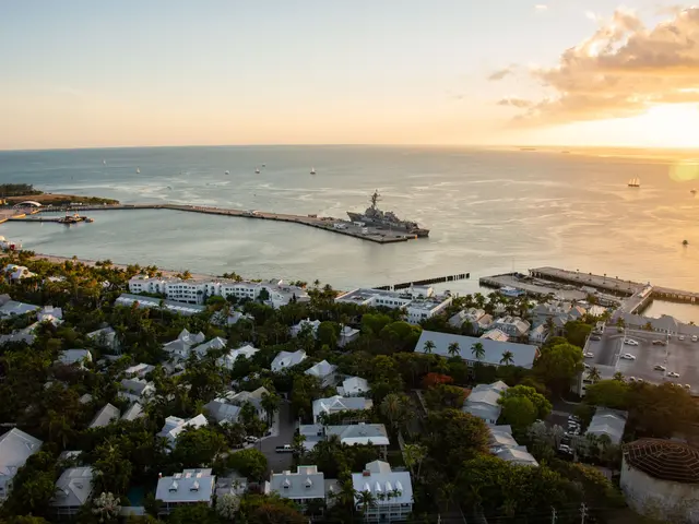 The image shows an aerial view of a city with boats in the water, surrounded by buildings, trees,...
