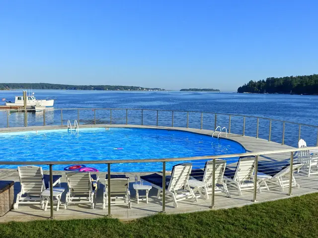 The image shows a large swimming pool with lounge chairs and a hot tub on the shore of a lake,...