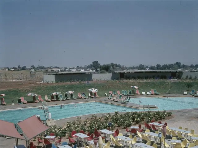 The image shows an outdoor swimming pool with tables and chairs around it, surrounded by plants...